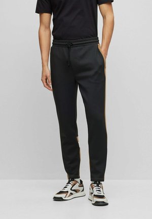 Hombre vistiendo pantalones jogger negros con franjas laterales marrones, camiseta negra y zapatillas blancas con detalles en negro y marrón, de pie contra un fondo liso.