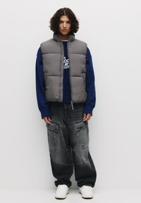 Puffet grå vest over en blå sweater, parret med løse sorte denim cargo bukser. Hvide sneakers fuldender outfittet.