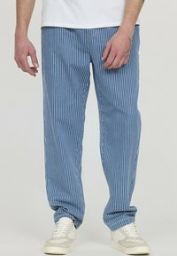 Pantalons rayés bleus en tissu léger, avec des rayures blanches verticales, des poignets élastiques et des poches latérales. Associés à des chaussures blanches.