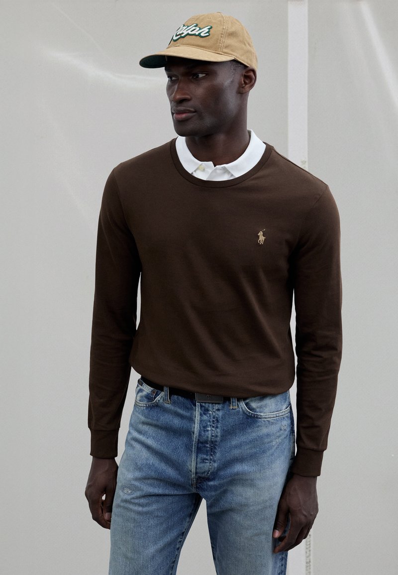 Brown long-sleeve shirt with a round neckline, layered over a white collared shirt. Light blue jeans; beige baseball cap with green text.