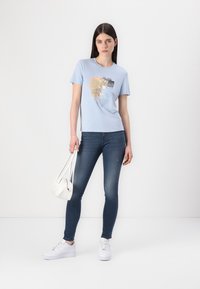 Lichtblauw T-shirt met korte mouwen en een kleurrijke grafische print. Gecombineerd met donkere skinny jeans en witte sneakers. Witte schoudertas.