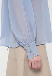 Mouw van een doorschijnende lichtblauwe blouse met dubbele knoopmanchet, over een beige leren rok, hand rustend naast het lichaam.