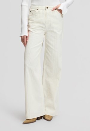 Femme portant un pantalon large en velours côtelé couleur crème à taille haute et un haut blanc côtelé à manches longues, debout avec une main dans la poche.