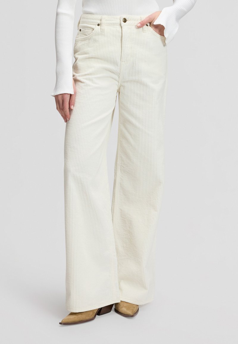Femme portant un pantalon large en velours côtelé couleur crème à taille haute et un haut blanc côtelé à manches longues, debout avec une main dans la poche.