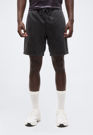 Mann trägt schwarze Shorts mit Kordelzug, weiße Sportsocken und weiße Sportschuhe, steht vor einem einfarbigen Hintergrund.