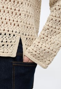 Χέρι σε τσέπη σκούρων μπλε τζιν, φορώντας μπεζ πλεκτό πουλόβερ με ανοιχτό μοτίβο και πλαϊνή σχισμή.