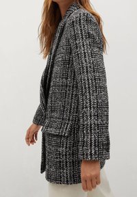 Blazer pied-de-poule noir et blanc en tissu texturé. Présente une coupe ample, de grandes poches et des boutons décoratifs au niveau du poignet.