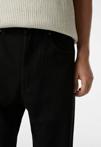 Pantalones de mezclilla negros de corte holgado, con costuras visibles, bolsillos laterales y cierre de botón. Material de algodón con ligera textura.