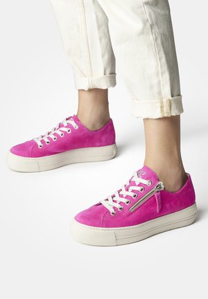 Sneaker in suede rosa con lacci bianchi e suola in piattaforma. Presenta un dettaglio con zip sul lato e una punta arrotondata. Pantaloni chiari arrotolati.