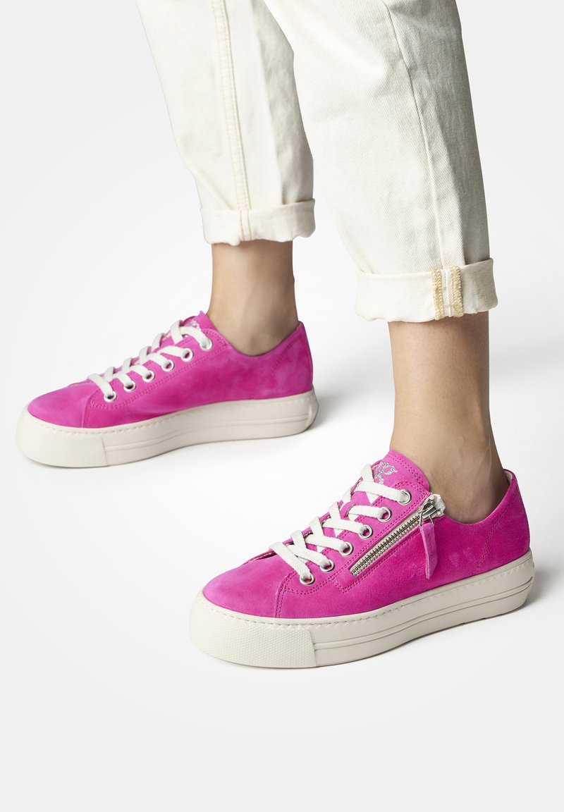 Sneaker in suede rosa con lacci bianchi e suola in piattaforma. Presenta un dettaglio con zip sul lato e una punta arrotondata. Pantaloni chiari arrotolati.