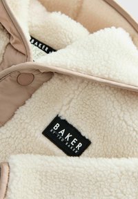 Beež jakk, millel on kreemjas, pehme vooder ning must silt valge tekstiga "BAKER by TED BAKER." Sisaldab nupu detaili.