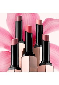 Cinq rouges à lèvres roses et nude avec des boîtiers noirs et beiges élégants, disposés sur un fond floral rose doux.