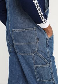 Pantalones de mezclilla azules con múltiples bolsillos, presentando un diseño de pierna recta y detalles de costura. La tela parece suave con un acabado desgastado.
