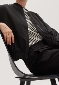 Czarna kurtka typu bomber z zamkiem błyskawicznym, wyposażona w elastyczne mankiety. Pod spodem koszula w paski z poziomymi liniami. Czarne spodnie i krzesło.