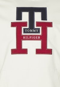Sudadera blanca con un logo bordado: una gran "H" roja con una "T" azul marino, y el texto "TOMMY HILFIGER" en negro debajo, sobre una tela suave.
