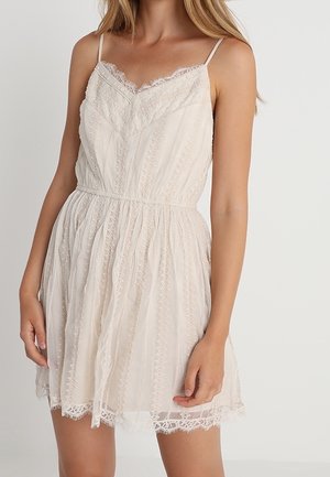 Frau trägt ein ärmelloses, knielanges Kleid aus cremefarbener Spitze mit dünnen Trägern und tailliertem Schnitt, steht vor einem einfarbigen hellen Hintergrund.