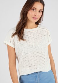 Top blanco de manga corta hecho de tejido perforado con patrones circulares. Presenta un escote redondeado y un corte relajado, combinado con vaqueros azul claro.