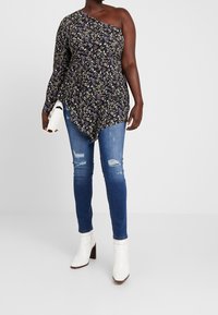 Top negro de flores con un hombro descubierto y dobladillo con volantes, combinado con jeans skinny azules desgastados y botas blancas de tobillo con tacón cuadrado.