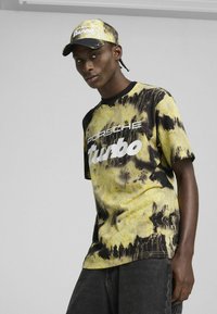 Žlté a čierne tričko s tie-dye motívom má biely nápis "Porsche Turbo". Je kombinované s tmavými džínsami a zodpovedajúcou čiapkou.