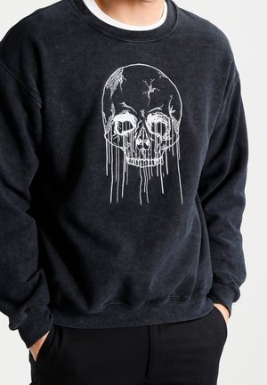 Sudadera negra con un gráfico de calavera blanca agrietada con líneas que gotean en el pecho, usada por una persona con las manos en los bolsillos.