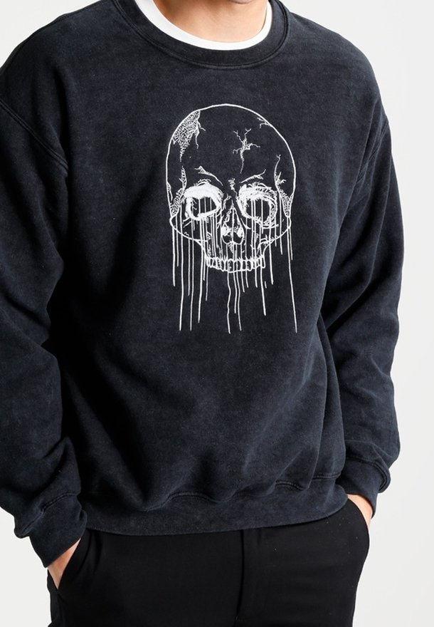 Sweat-shirt noir avec un motif de crâne blanc fissuré avec des lignes dégoulinantes sur la poitrine, porté par une personne les mains dans les poches.
