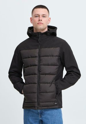 Young man with short hair wearing a black puffer jacket with a hood, standing with hands in pockets against a plain light background.