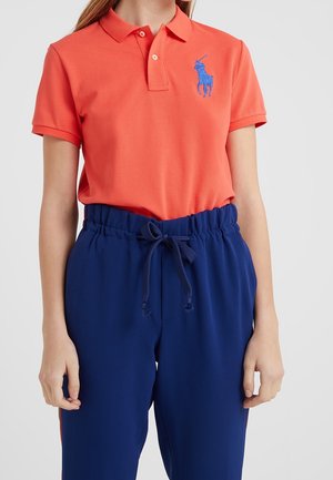 Oranges Poloshirt mit blau besticktem Logo, kombiniert mit marineblauen Hosen mit Kordelzug. Der Stoff wirkt glatt und der Schnitt ist entspannt.