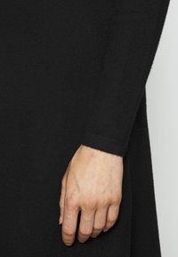 Vêtement en tricot noir à manches longues, avec une zone ajustée au poignet. Une main visible repose sur le tissu, illustrant une texture lisse.