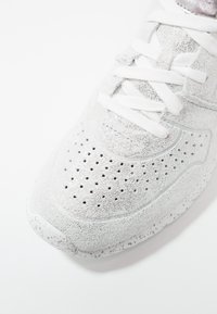 Baskets blanches avec embout perforé, dessus texturé et lacets blancs sur fond blanc.