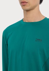 Langarm teal sportshirt gemaakt van een gestructureerde stof, met een ronde halslijn en een klein logo op de linkerkant van de borst.