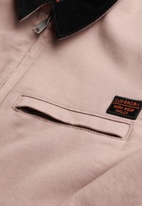 Close-up af pink stofjakke med sort krave, lynlåslomme på brystet og rektangulært sort og orange "Superdry Work Wear" mærke.