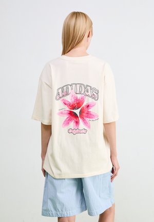 Personne aux longs cheveux blonds vue de dos, portant un t-shirt crème Adidas avec un grand motif de fleur rose et un short bleu clair à rayures.