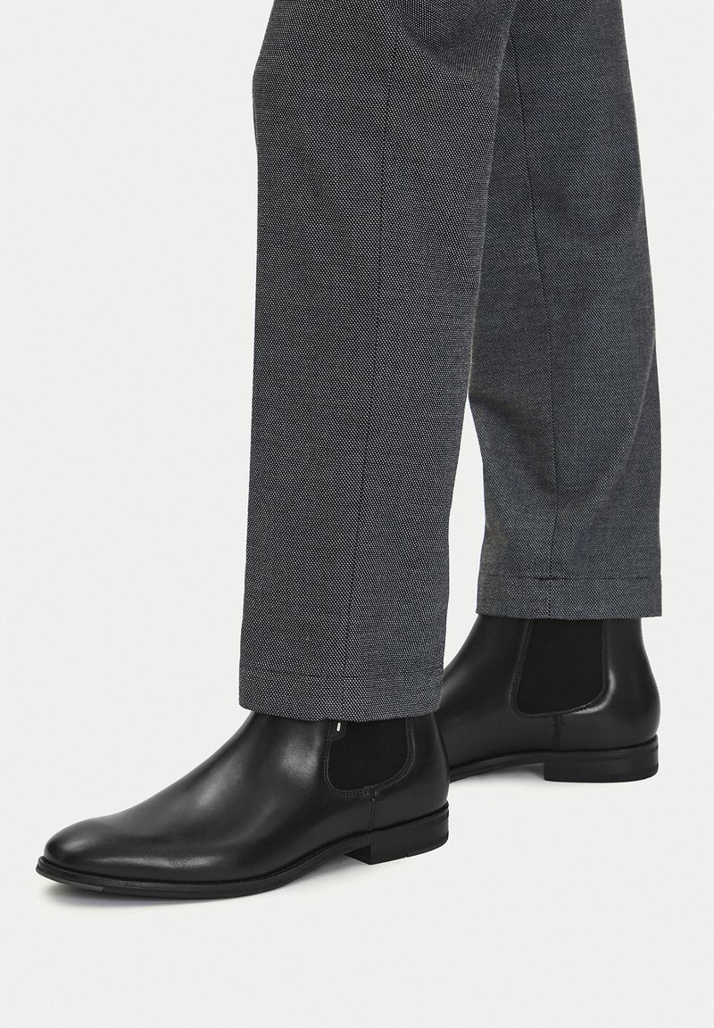 Crne kožne gležnjače s elastičnim bočnim panelima, zaobljenim vrhom i niskom petom, uparene sa sivim teksturiranim hlačama koje se sužavaju kod gležnja.