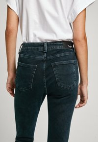 Jeans de mezclilla negra con un corte ajustado, que cuentan con dos bolsillos traseros y una etiqueta de cuero de marca en la cinturilla. Textura suave, poco desgaste.