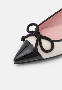 Chaussure plate à bout pointu avec embout en cuir verni noir, corps crème, intérieur rose et détail de nœud en corde noire torsadée sur le dessus.