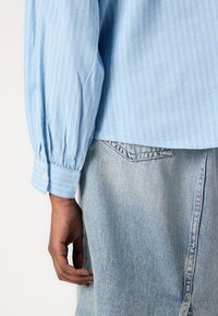 Camisa riscada azul clara com punhos abotoados, combinada com calças de ganga claras. O tecido parece macio e tem um ajuste descontraído.
