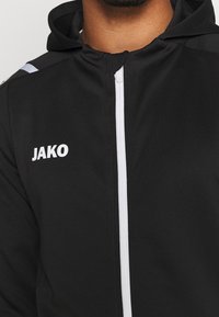 Svart Jako sportjacka med vit dragkedja, utrustad med huva och texturerat tyg på axlarna, med varumärkeslogotypen i vitt.