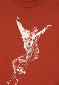 T-shirt couleur rouille arborant un graphique blanc d'un skateur en mouvement, avec des textures détaillées dans les vêtements et les éléments du skateboard.
