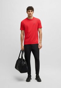 Rød kortærmet sports skjorte med rund hals og et lille sort logo, parret med sorte slim-fit bukser og en sort duffel bag.