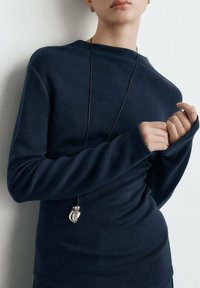 Maglione blu navy con vestibilità comoda, tessuto strutturato, maniche lunghe e collo alto. Un ciondolo d'argento pende da un cordoncino nero.