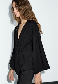 Jeune femme aux cheveux longs et ondulés porte une veste noire ample avec des manches larges et un décolleté en V plongeant, se tenant devant un fond uniforme.