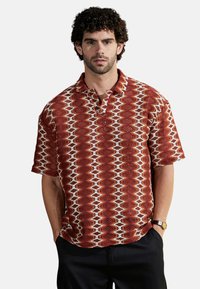 Camisa de manga corta con cuello en rojo, con un patrón geométrico, textura tejida y acentos en blanco. Combinada con pantalones oscuros y un reloj.