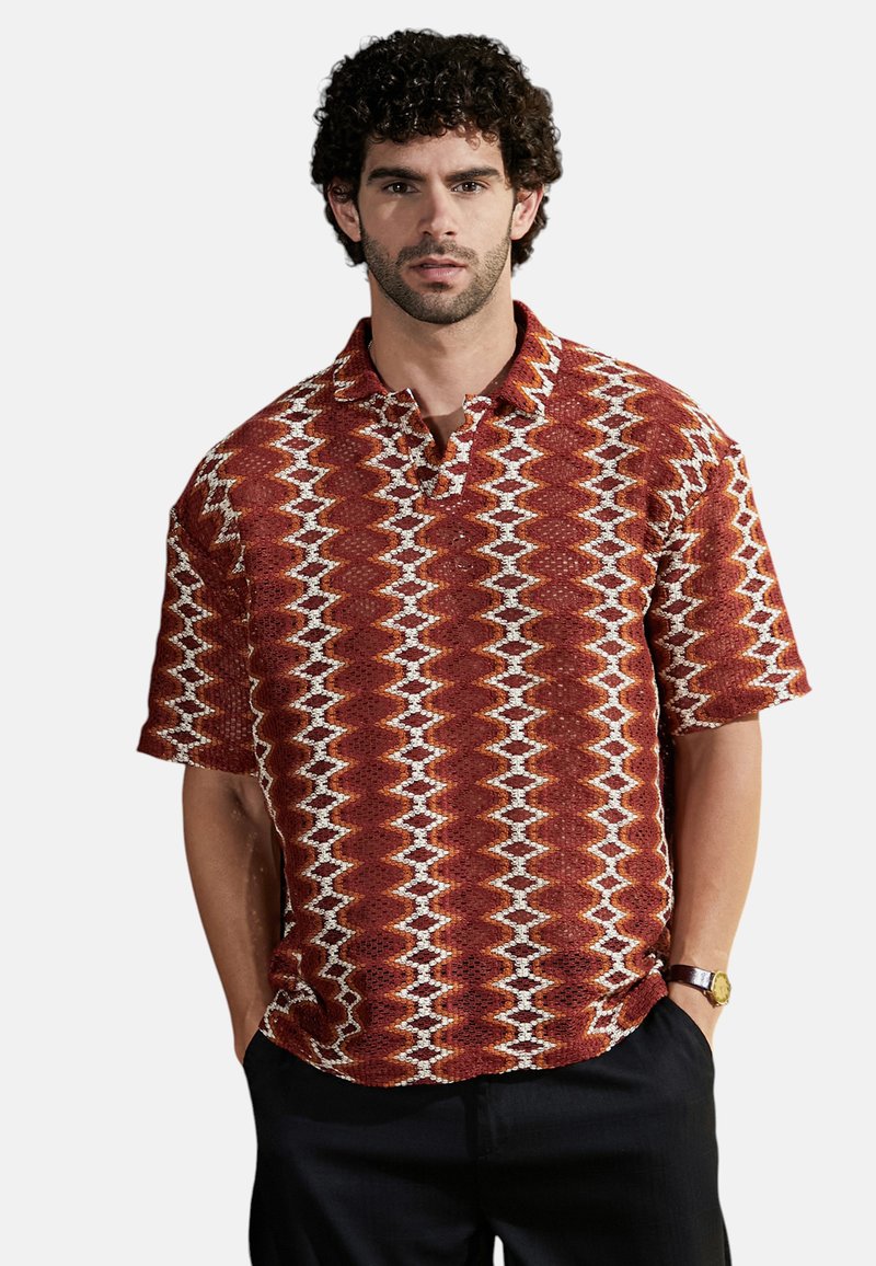 Camisa de manga corta con cuello en rojo, con un patrón geométrico, textura tejida y acentos en blanco. Combinada con pantalones oscuros y un reloj.