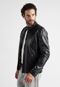 Schwarze Lederjacke mit Stehkragen, Reißverschluss vorne, zwei Brusttaschen und glatter Textur. Tragen über einem weißen Hemd und hellen Jeans.