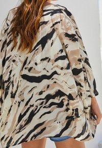 Lekki, przezroczysty kimono z abstrakcyjnym beżowo-czarnym wzorem zwierzęcym. Posiada luźne rękawy i swobodny krój, podkreślający ziemistą paletę kolorów.