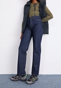 Dark blue denim jeans with a straight leg fit. Paired with a green tie-dye long-sleeve top and a dark gray puffer vest. Black athletic shoes.