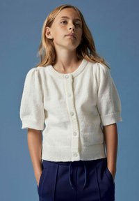 Fille avec de longs cheveux blonds portant un cardigan en tricot à manches courtes blanc avec des boutons en forme de fleur et un pantalon bleu marine, sur un fond bleu.