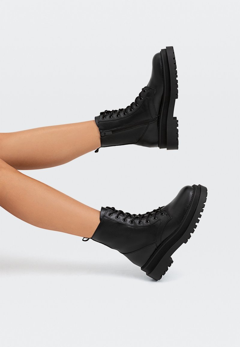 Jambes portant des bottes de combat noires à lacets avec semelles épaisses, levées horizontalement sur un fond gris neutre.