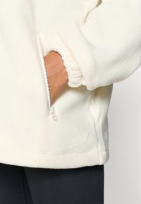 Valkoinen fleecetakki, jossa vetoketjullinen tasku ja joustavat hihansuut, pehmeällä, sileällä tekstuurilla ja rennolla istuvuudella.