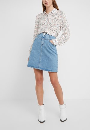 Femme portant un chemisier blanc à pois multicolores, une jupe en jean bleue et des bottines blanches, debout avec une main dans la poche.